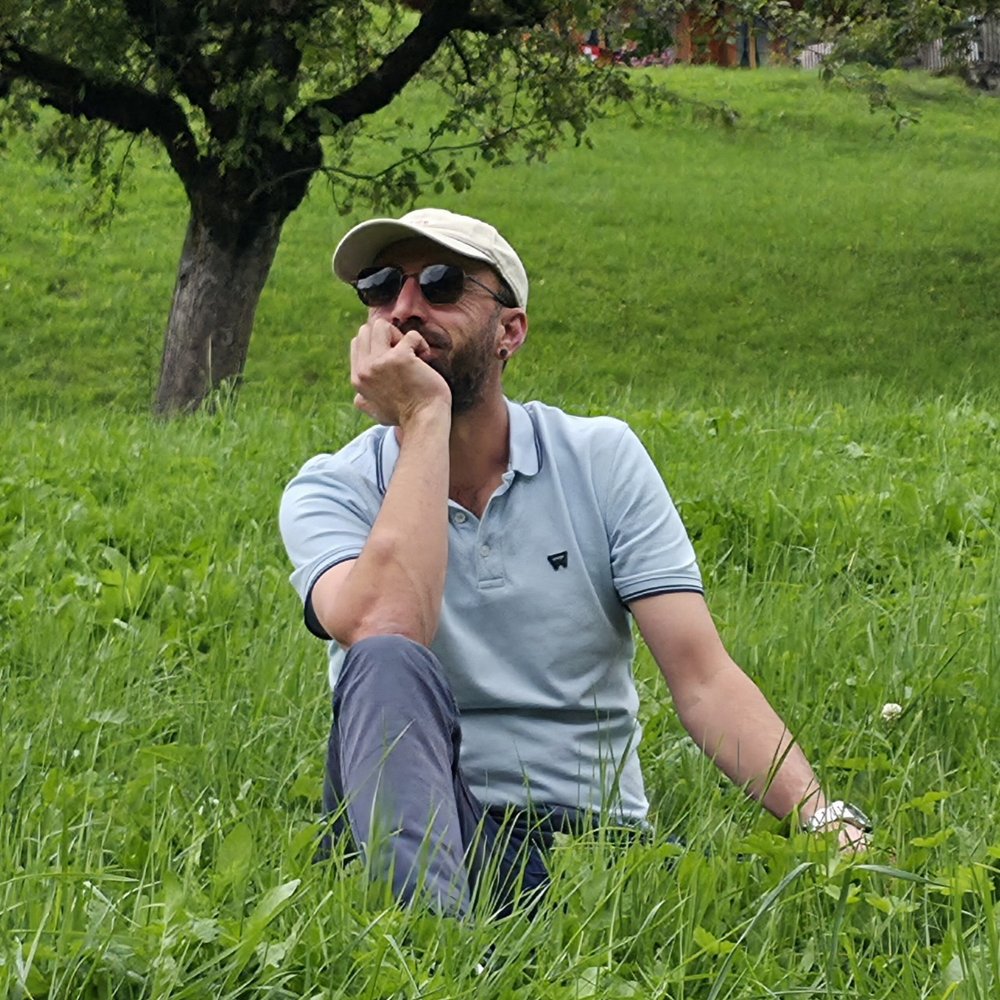 halit yesil otlarin uzerinde oturuyor, arkada ahsap isvicre evi ve bir agaç, mavi gökyüzü